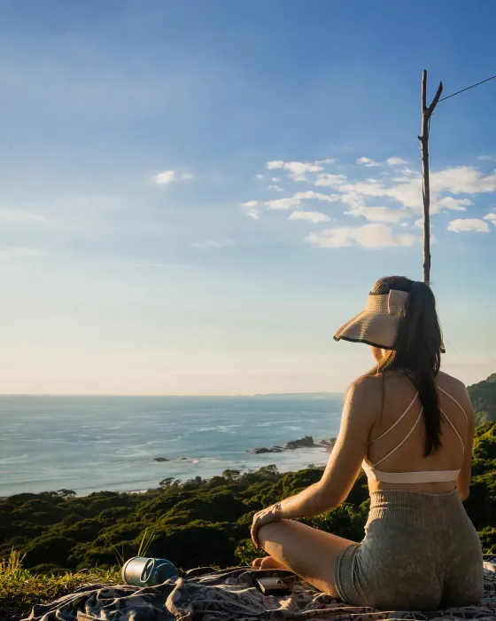 A woman sitting cross-legged on the ridge at Amora in late afternoon light, seen from behind, looking out over the Costa Ballena coastline far below