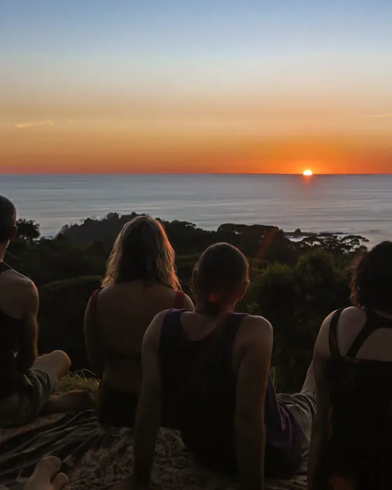 People sitting shoulder to shoulder on the ground at dusk, watching the sun reach the horizon