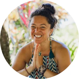 Ariana Binney, Steward of Education, smiling with her hands pressed together in greeting, outdoors among tropical plants.