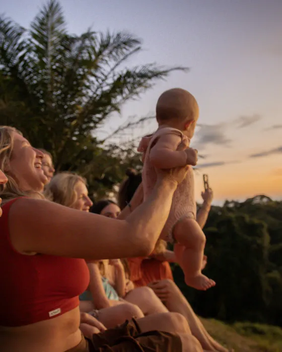 A woman lifting a laughing baby up into the last of the light while others nearby watch the dusk sky
