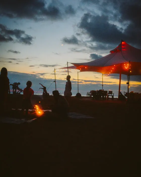 The first fire of the evening beside the lantern-strung tent at Amora at dusk, a crescent moon over the Pacific