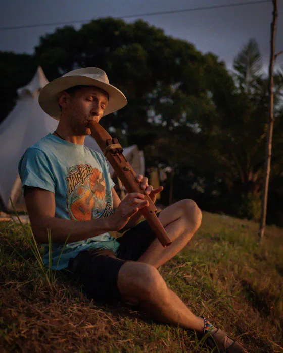 A man in a sun hat playing a wooden flute on the open hillside at Amora as the light goes