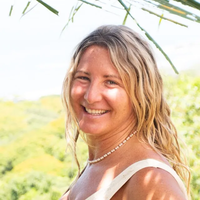 Jessica Filkins, Visionary Director of Amora, smiling outdoors under a palm frond with green forest behind her.