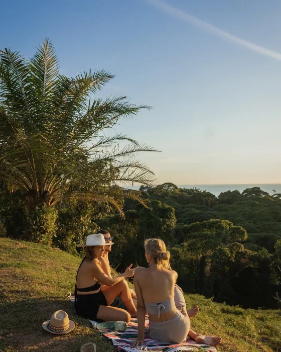 Three women sitting on a picnic blanket under a palm tree at Amora in warm late light, the Pacific in the distance behind them