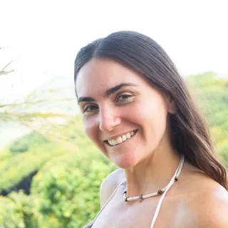 Victoria Leyden, Steward of Community, smiling outdoors wearing a wooden bead necklace, a green hillside behind her.