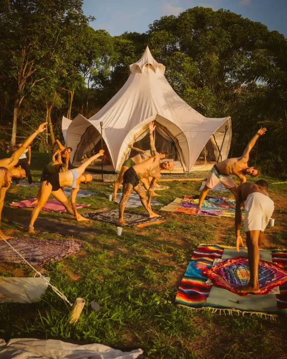 The community stretching side to side on the grass in front of the lotus bell tent at Amora, late sun on the trees — a yoga session on the land of an ocean-view ecovillage in Costa Rica