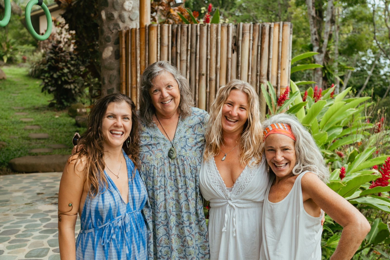 Four women standing arm in arm on the stone path, laughing, bamboo and red ginger flowers behind them.