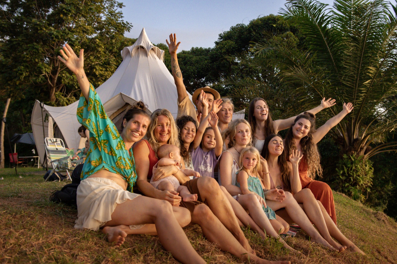 Women and children sitting close together on the grass in front of a bell tent, arms raised in the last golden light.